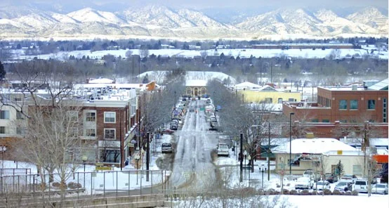 Snow-covered street lined with buildings and parked cars in Littleton, Colorado, with leafless trees and mountains in the background under a cloudy sky.