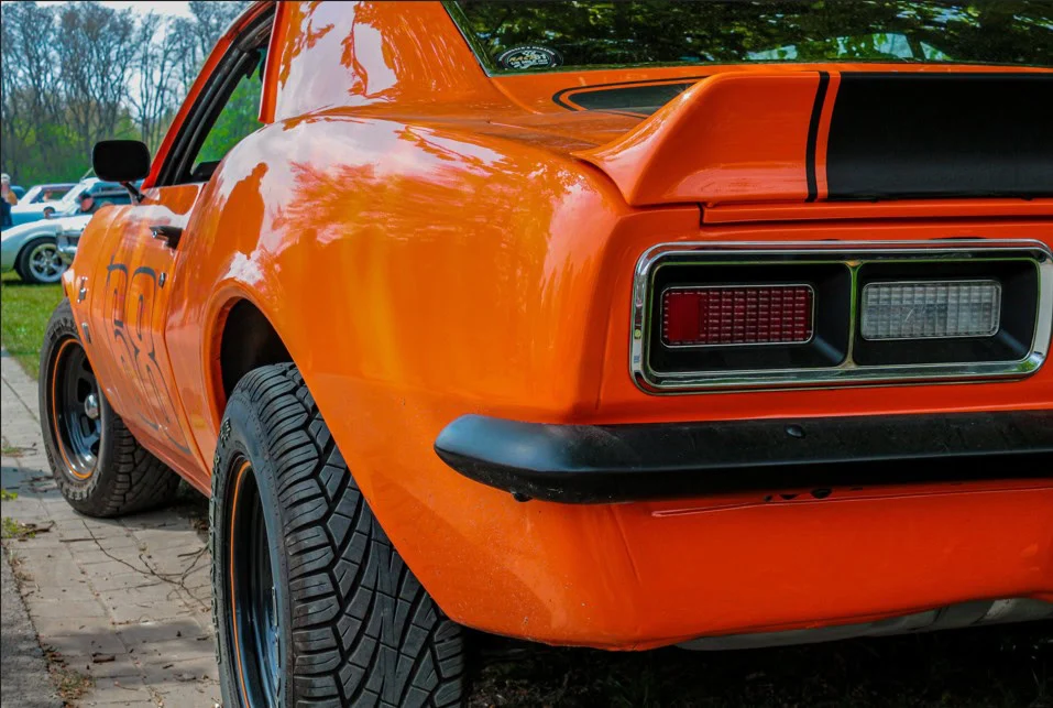 Close-up of the rear left side of an orange classic car with black detailing, parked on a paved surface, with trees and other cars in the background—a perfect scene for enthusiasts who appreciate expert vehicle maintenance like that offered at Grease Monkey Rexburg.