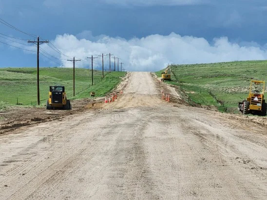 Unpaved rural road under construction with construction vehicles, traffic cones, and power lines, set in a grassy Elizabeth Colorado landscape under a cloudy sky.