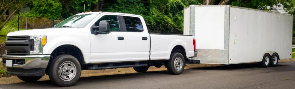A white pickup truck is parked on a street in Pocatello, Idaho, towing a large enclosed white trailer with two axles. Trees and a fence are visible in the background, suggesting the vehicle may be ready for travel after a recent brake system service at Grease Monkey.