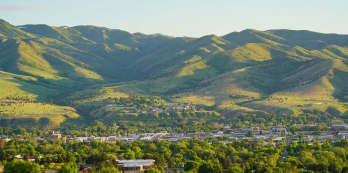 A town with many trees in the foreground sits below green, rolling hills under clear daylight, reminiscent of picturesque landscapes found around Pocatello, Idaho.