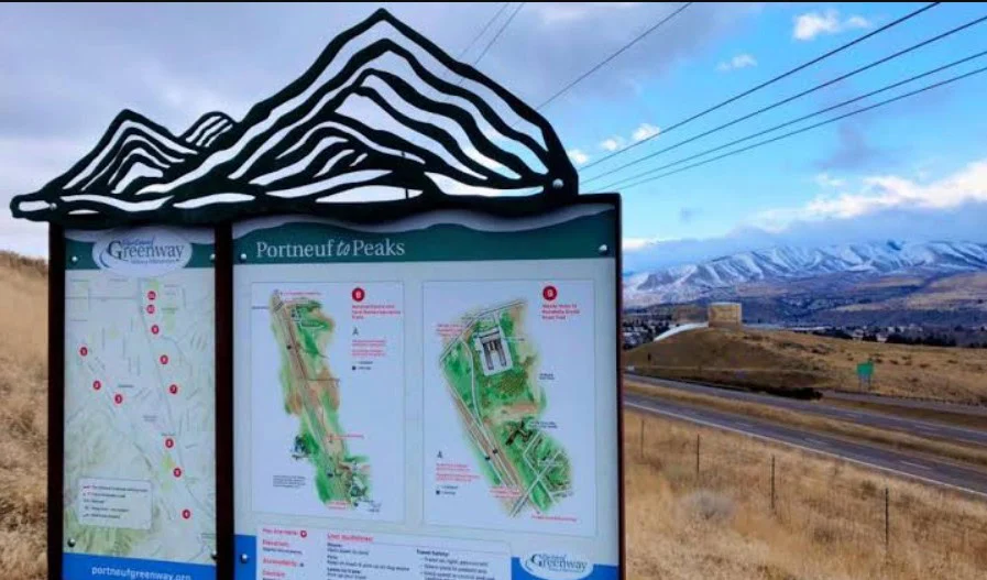 Trailhead sign for Portneuf to Peaks Greenway stands beside a road in West Pocatello, with mountains and grassy hills showcasing the region’s unique bench-to-canyon geography under a partly cloudy sky.