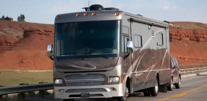 A large gray motorhome is driving on a road through the desert landscape of Magic Valley, towing a smaller brown vehicle behind it.