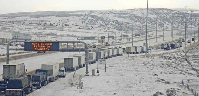 A long line of trucks waits on a snow-covered highway near Laramie, Wyoming, next to a sign reading “ROAD CLOSED RETURN TO EVANSTON.” Snow blankets the ground and hills in the background, as drivers likely consider important safety measures such as brake inspection or brake service before continuing their journey.