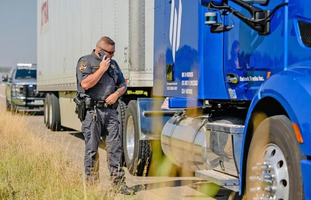 A police officer stands next to a blue semi-truck, discussing a possible brake system issue over the radio, with a police vehicle parked nearby on the roadside near Pocatello Idaho.