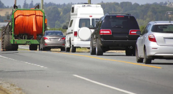 Several vehicles, including a tractor, RV, and cars—perhaps on their way to Jerome Idaho for a vehicle service—travel in a line on a two-lane rural road with trees and hills in the background, each carefully relying on their brake system as they navigate the scenic route.