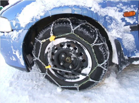 Close-up of a car tire fitted with snow chains on a snowy road near Fort Carson, attached to a blue vehicle’s wheel for improved traction and transmission performance in winter conditions.