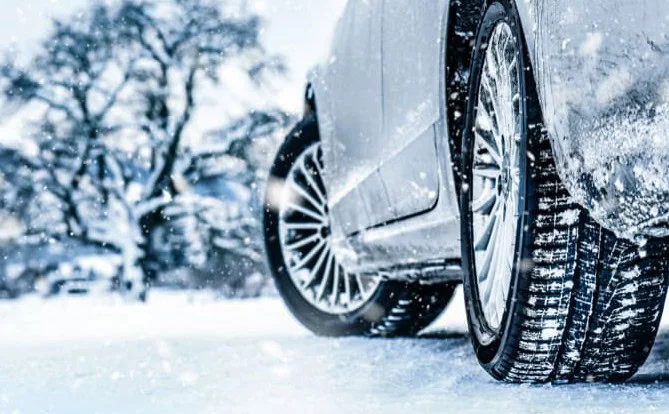 Close-up of a car’s tire on a snow-covered road near Kelly Canyon with trees in the background, showing winter driving conditions, falling snowflakes, and the importance of reliable transmission performance.