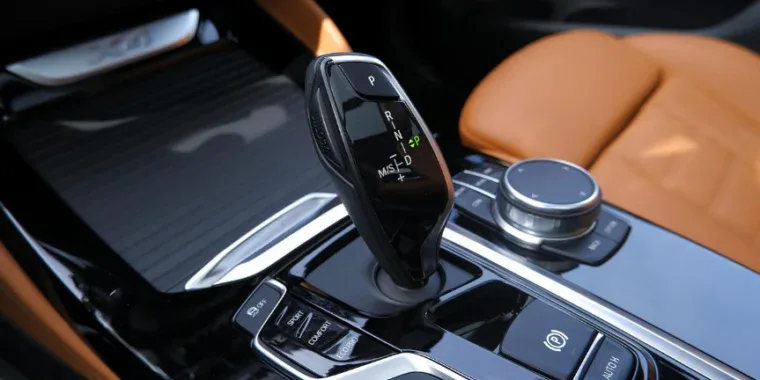 Close-up of a modern car’s center console featuring a black electronic transmission gear shifter, control dial, and tan leather seat.
