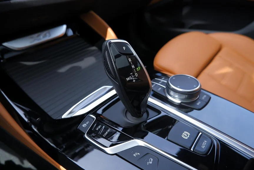 Close-up of a modern car’s center console featuring a black electronic transmission gear shifter, control dial, and tan leather seat.
