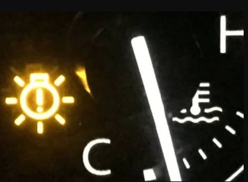 Close-up of a car dashboard showing a yellow warning light with a bulb and exclamation mark, next to a coolant temperature gauge—captured by 2nd East Transmission on the Business Loop in Rexburg.