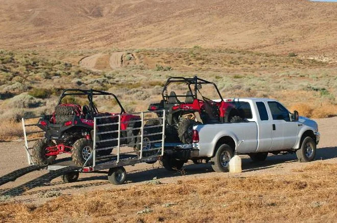 A white pickup truck is parked on a dirt road, towing a trailer with two red off-road vehicles in a dry, hilly landscape—perfectly maintained and ready for adventure thanks to regular vehicle maintenance at Grease Monkey Rexburg.