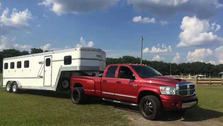 A red pickup truck is parked on grass, towing a large white horse trailer under a partly cloudy sky near Idaho Falls, highlighting the importance of regular vehicle maintenance and brake system service for safe travels.