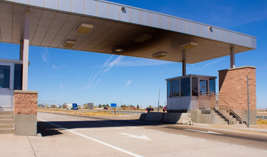 A toll booth with two small buildings stands over an empty road under a clear blue sky, set against the backdrop of Jerome, Idaho’s sprawling dairy country, with distant signs and buildings visible in the background.