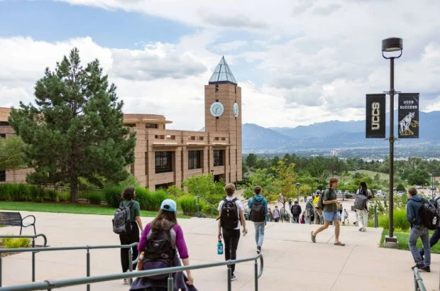 Students walk on a campus path toward a building with a clock tower in Colorado Springs; UCCS banners and mountains are visible in the background.
