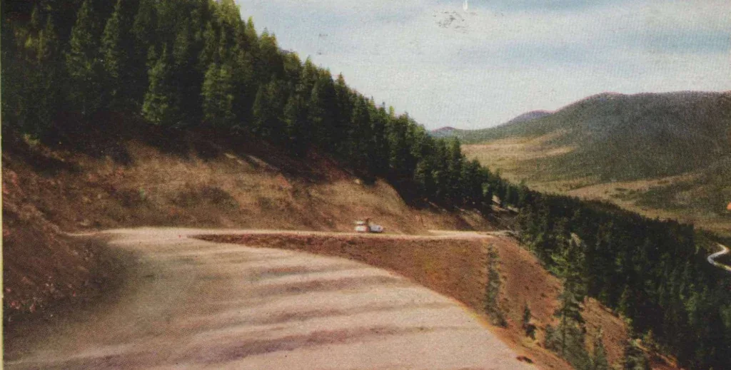 A winding mountain road curves around a steep slope lined with dense trees and distant hills under a blue sky, offering Ute Pass drivers breathtaking views as they approach Woodland Park.