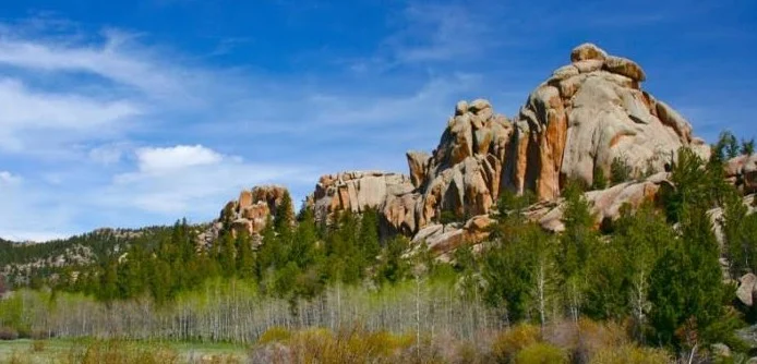 Rocky formations rise above a landscape of green trees and shrubs under a blue sky with scattered clouds, creating a scenic view reminiscent of the rugged beauty found near Laramie, Wyoming.