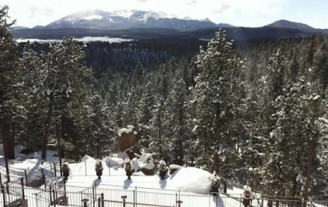 Snow-covered trees and a fenced area in the foreground evoke the serene beauty of Woodland Park, with Ute Pass rising in the distance beyond mountains and a shimmering lake under a partly cloudy sky.