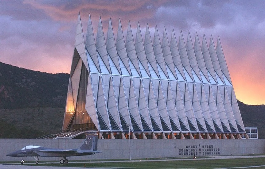 The U.S. Air Force Academy Cadet Chapel, located in Colorado Springs, stands majestically with a jet parked in front, set against a dramatic sunset sky and the towering mountains in the background.