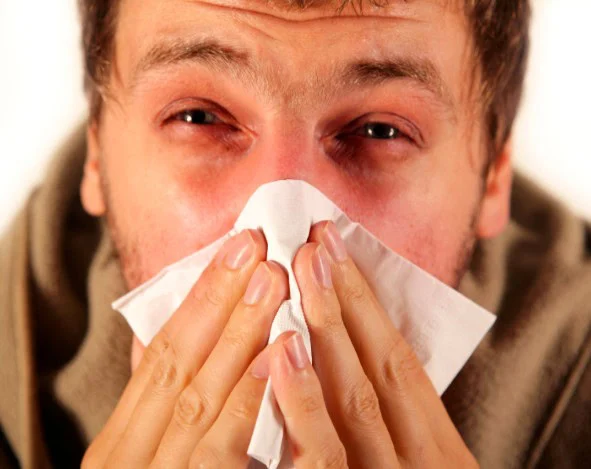 A person holds a tissue to their nose and appears to be sneezing or blowing their nose, with red, watery eyes and a flushed nose—possibly suffering from allergies that could be eased by regular car maintenance such as replacing the cabin air filter.