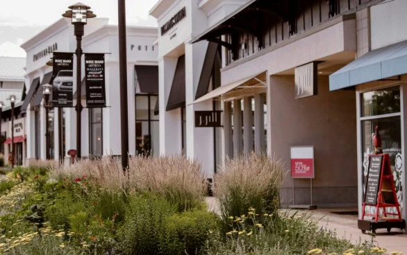 Outdoor shopping center with storefronts, sidewalk, and landscaped greenery in front; signs and advertisements visible along walkway—perfect for browsing shops or attending to car maintenance like replacing a cabin air filter in Littleton, Colorado.