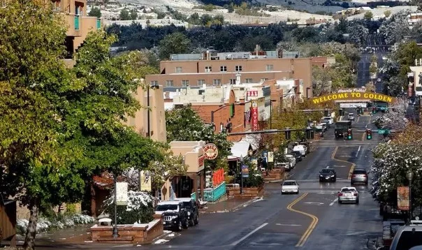 Downtown street in Golden, Colorado with cars, shops, trees, and a yellow “Welcome to Golden” archway over the road; snow is visible on rooftops and greenery.