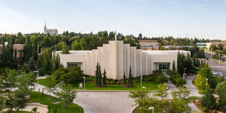 A modern, white, stepped building sits amid green trees and landscaped lawns in Rexburg Idaho, with a road and more buildings in the background under a partly cloudy sky.