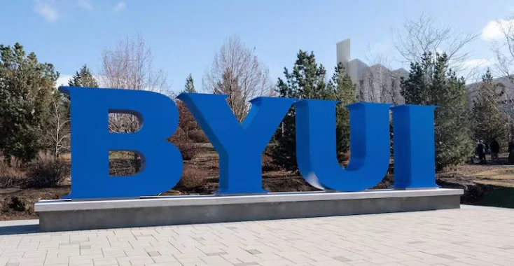 Large blue letters “BYUI” stand on a sidewalk in Rexburg, Idaho, with trees and buildings in the background under a clear sky.