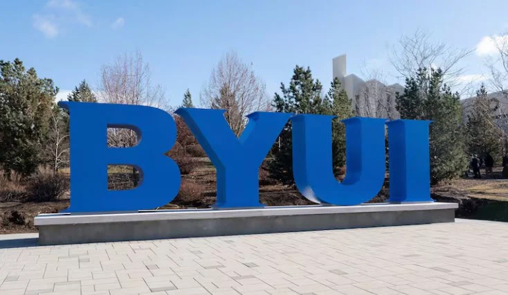 Large blue letters “BYUI” stand on a sidewalk in Rexburg, Idaho, with trees and buildings in the background under a clear sky.