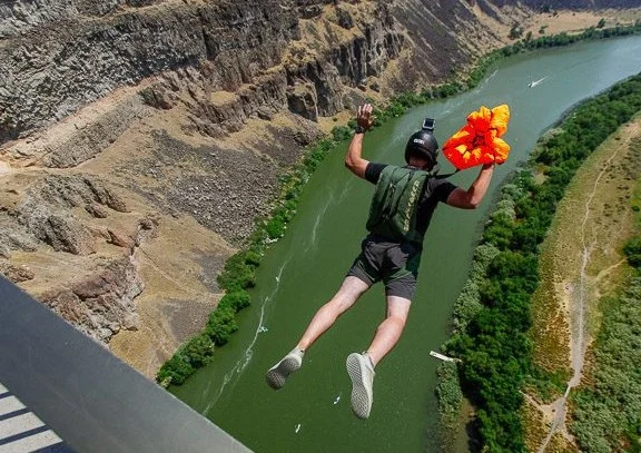 A person jumps off a bridge above a river in Twin Falls, Idaho, holding a parachute in hand, with a rocky canyon and lush greenery visible below.