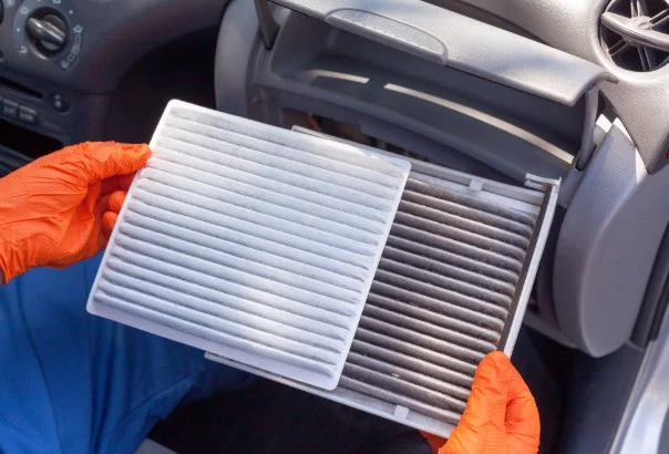 A person wearing orange gloves holds a clean cabin air filter next to a dirty one inside a car, demonstrating the difference in color and cleanliness—a clear reminder of the importance of regular car maintenance, especially for drivers in Rexburg Idaho.