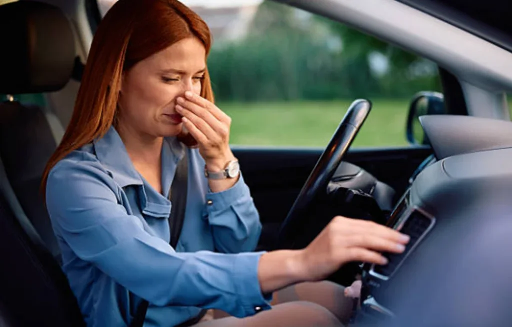 Woman sitting in a car in Rexburg Idaho holds her nose with one hand and adjusts the air conditioning controls with the other, appearing bothered by an unpleasant smell—an issue that could be solved with regular car maintenance, such as replacing the cabin air filter.