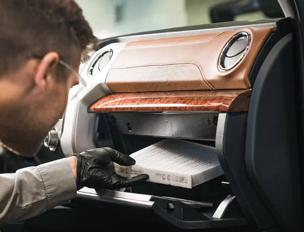 A person wearing gloves installs a cabin air filter into the glove compartment of a car dashboard with brown and wood accents, preparing for a smooth drive along Highway 30 near Pocatello, Idaho.