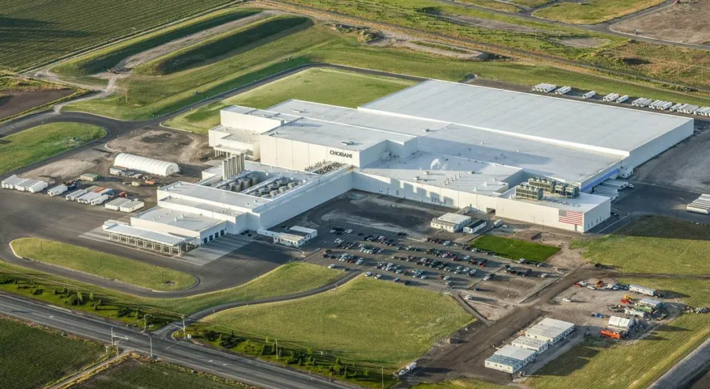 Aerial view of a large industrial facility with a white roof, parking lot with cars, and surrounding green fields and roads in Twin Falls, Idaho.