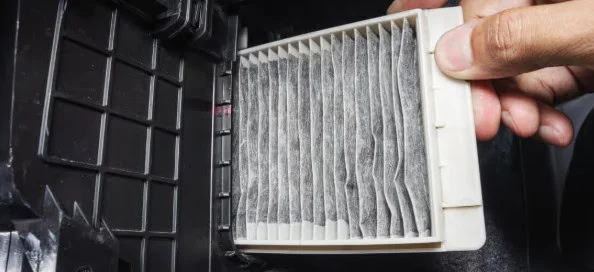 A hand is removing a dirty rectangular cabin air filter from a black compartment, likely in a vehicle or appliance, possibly near Aurora, Colorado or Buckley Space Force Base.