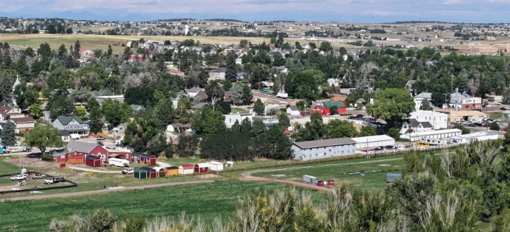 A small town in Elizabeth, Colorado, with houses, buildings, and trees is surrounded by open fields and distant hills under a partly cloudy sky.