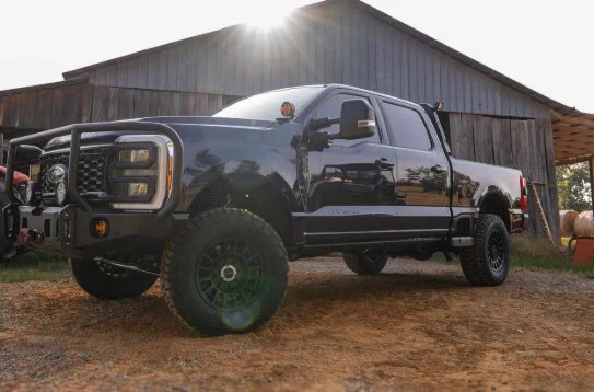 A black lifted pickup truck with off-road tires is parked on a dirt surface in front of a wooden barn, the sun shining above the roof—a classic scene from Kimberly, Idaho, capturing the spirit of Magic Valley farming.
