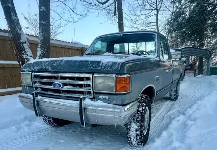 A gray Ford pickup truck with snow on its hood and tires is parked on a snowy driveway beside a wooden fence and trees, not far from Highway 30 in Pocatello West Side.