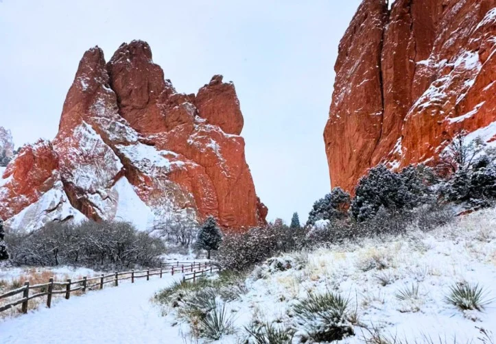 Snow covers the ground and red rock formations at Garden of the Gods park in Colorado Springs, with a wooden fence lining the snowy walking path under a cloudy sky.