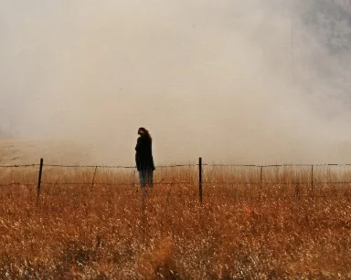 A person stands alone in a dry, grassy field near a wire fence with heavy smoke or fog in the background, just outside Buckley Space Force Base in Aurora, Colorado.