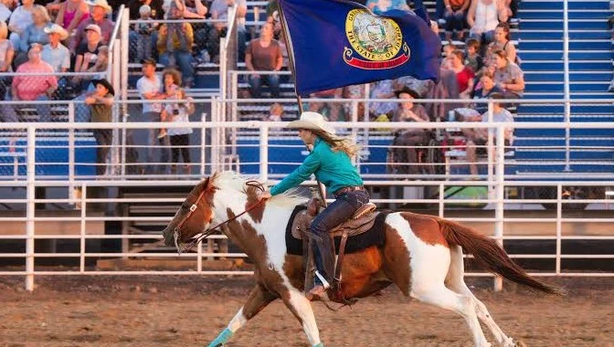 A woman rides a brown and white horse while waving a blue Idaho state flag in front of a crowd seated on blue bleachers at a rodeo event in Jerome, Idaho.
