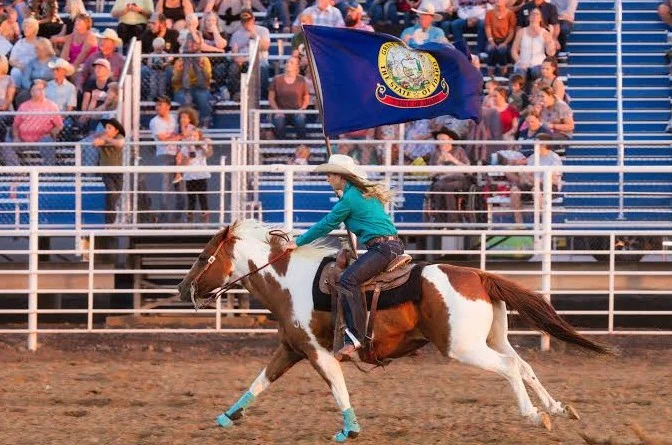 A woman rides a brown and white horse while waving a blue Idaho state flag in front of a crowd seated on blue bleachers at a rodeo event in Jerome, Idaho.