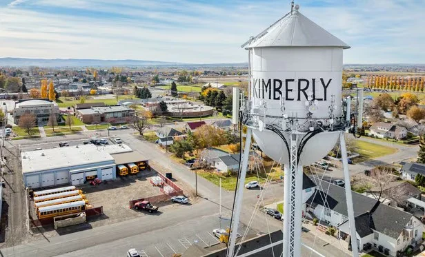 A white water tower labeled “Kimberly” stands above the small town of Kimberly, Idaho, with houses, school buses, and buildings visible below on a sunny day—nestled in the heart of Magic Valley farming country.