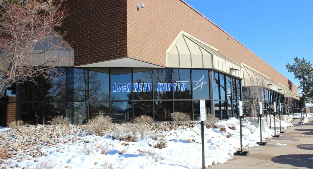 A brown brick building with large glass windows showing the Lockheed Martin logo stands in Littleton, Colorado; the surrounding area has snow and leafless shrubs, reminiscent of a winter day perfect for catching up on car maintenance such as replacing your cabin air filter.