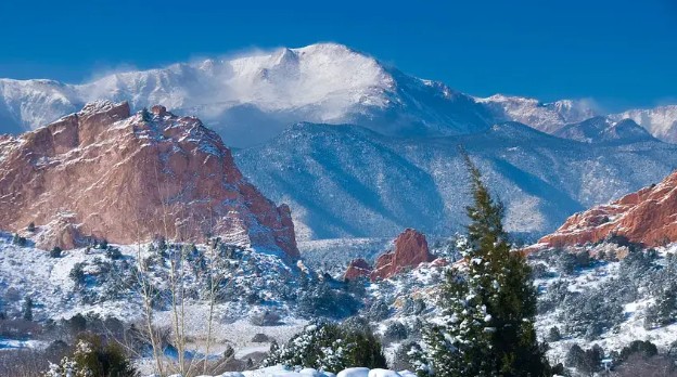 Snow-covered mountains rise behind red rock formations and evergreen trees under a clear blue sky, capturing the natural beauty surrounding Colorado Springs.