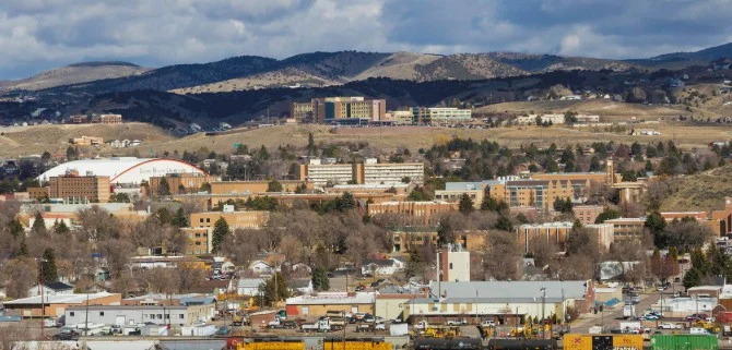 A small cityscape with a mix of residential, commercial, and institutional buildings—set against rolling hills just outside Pocatello, Idaho—unfolds beneath a partly cloudy sky, with Highway 30 winding through the scene.