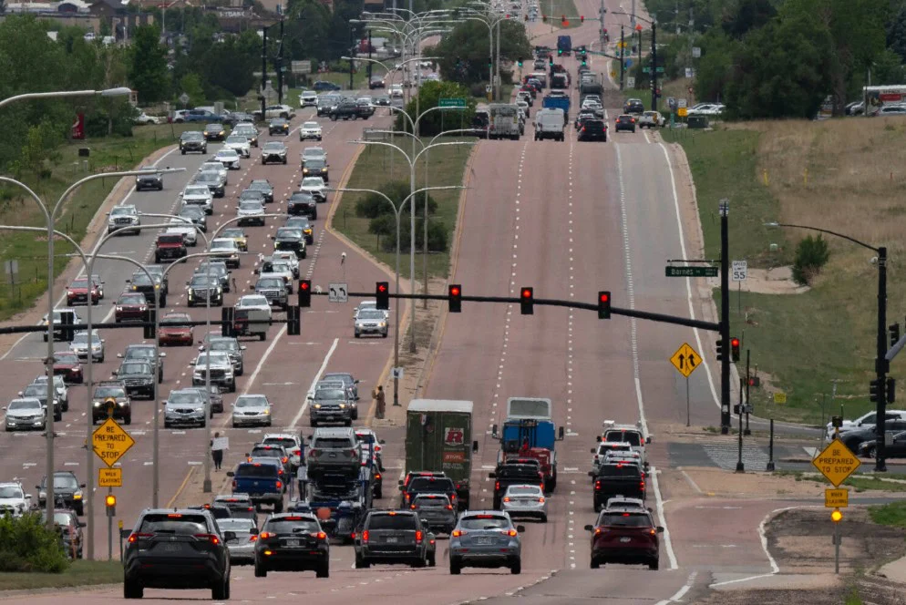 Heavy traffic fills multiple lanes at a red light on a divided road near Fort Carson, with cars stopped in both directions and some moving in the distance. Trees and grassy areas line the sides, reflecting the typical scenery found around Colorado Springs.