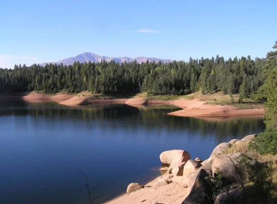 A calm lake with clear water is bordered by trees and sandy shores, with a distant forested mountain visible under a blue sky—evoking the serene landscapes found near Woodland Park, Colorado, in the heart of wildfire country.