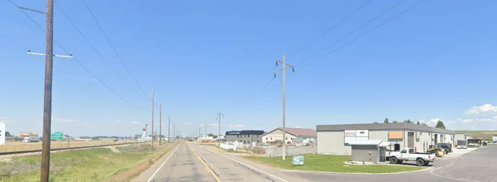 A rural road in Rexburg Idaho runs beside power lines and light industrial buildings under a clear blue sky, with parked vehicles—some possibly awaiting car maintenance or a new cabin air filter—visible near the buildings.