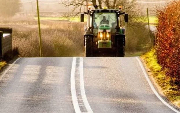 A green tractor drives on a rural paved road with double white lines, surrounded by fields and bushes on a sunny day near Rexburg, Idaho—a peaceful scene that highlights the importance of regular car maintenance, such as replacing your cabin air filter, for smooth travels through picturesque countryside.